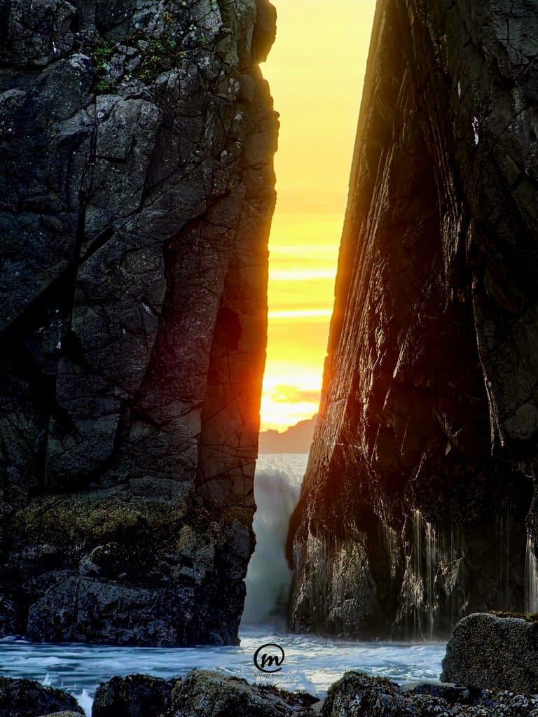 Alt Text Vibrant yellow sunset glowing through a narrow gap between two large, dark coastal sea stacks with crashing ocean waves.
