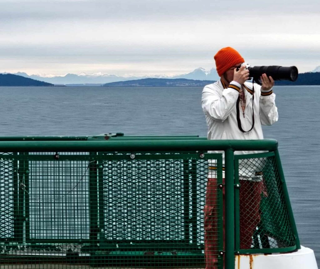 Photographer, Alexander Malloy in an orange beanie using a telephoto lens on a ferry deck with ocean and mountains in the background.