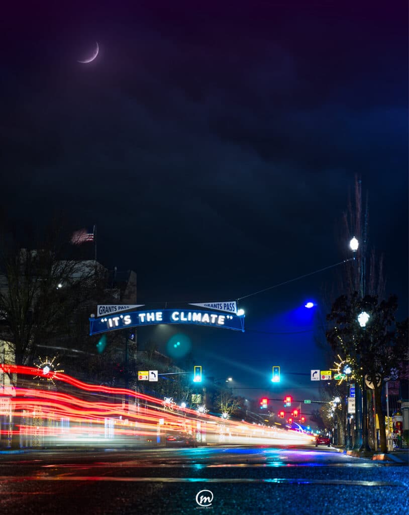 Nighttime street view in downtown Grants Pass with car light trails under the It's The Climate sign and a crescent moon.