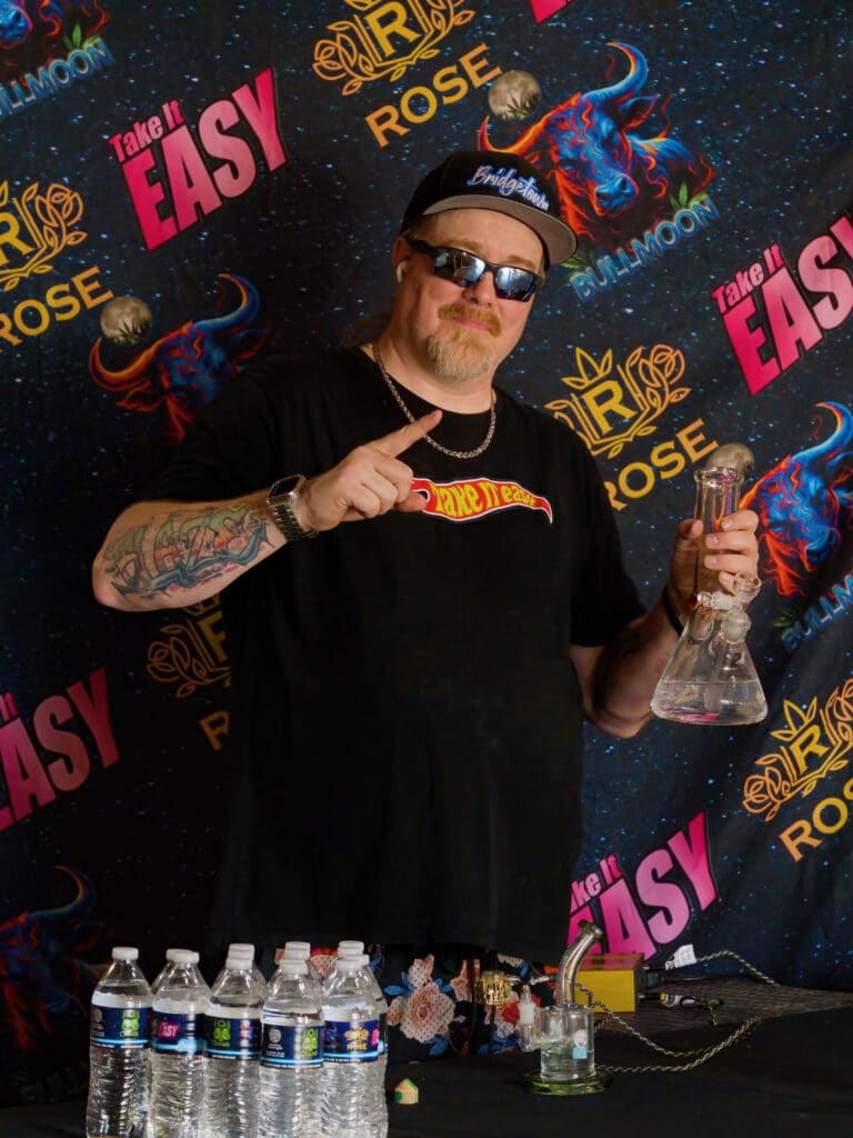 DJ Blaze wearing a black "Take It Easy" t-shirt gestures while holding a glass dab rig in front of a colorful indoor event backdrop