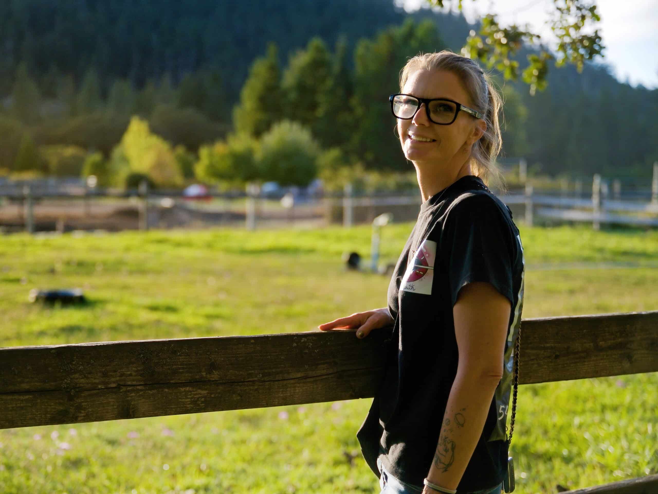 Vegi Hoyt of In Your Mouth LLC smiling by a rustic wooden fence in a sunny green pasture.