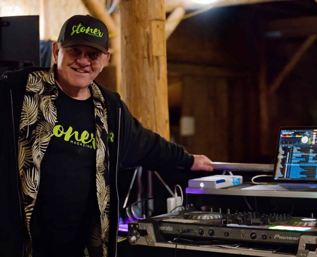 Jerry DeFoe wearing Stoner Magazine gear smiling next to a Pioneer DJ setup and laptop in a rustic wooden venue.