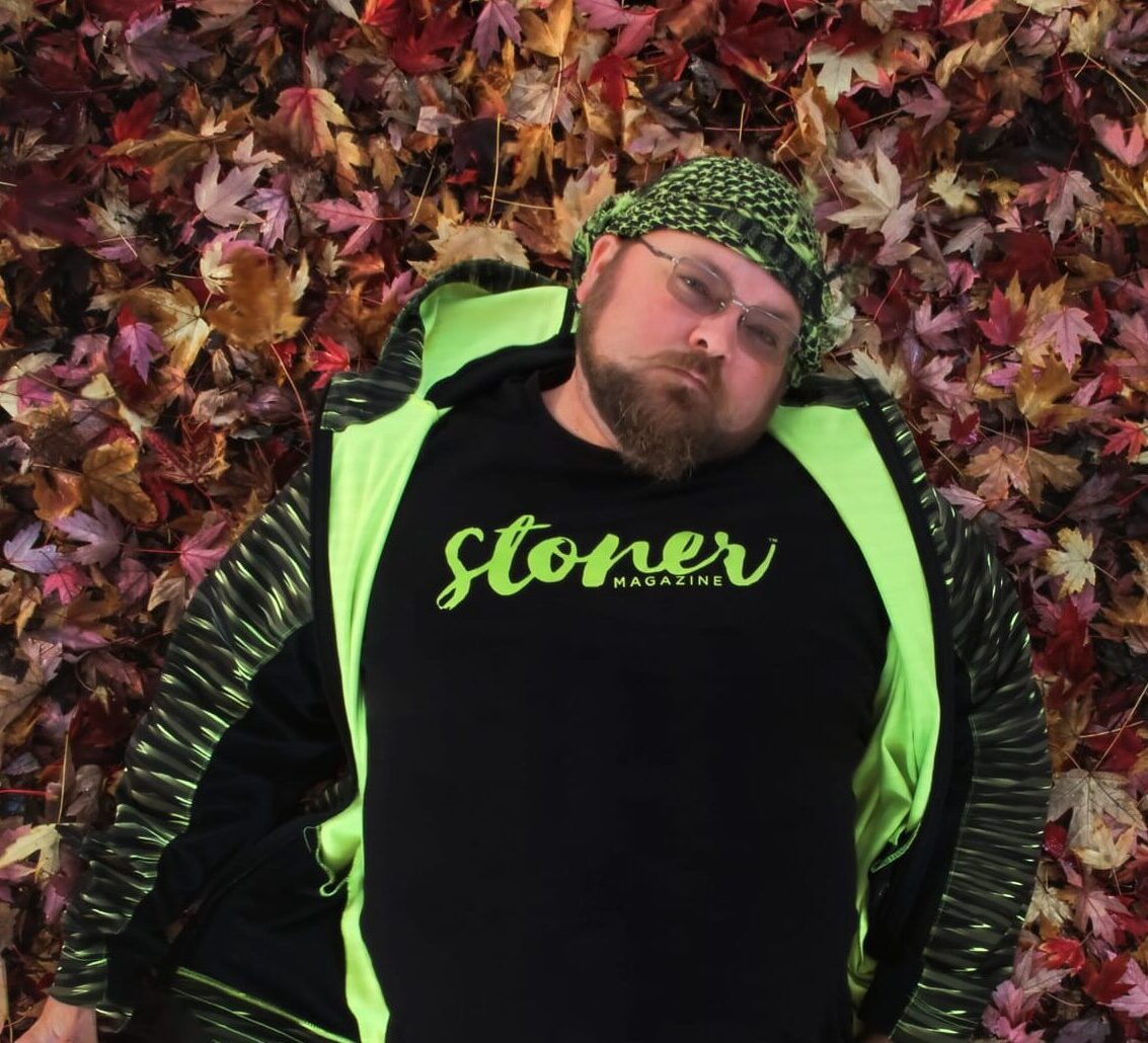 Man lying on colorful autumn leaves wearing a neon green jacket, beanie, and a black stoner magazine t-shirt.