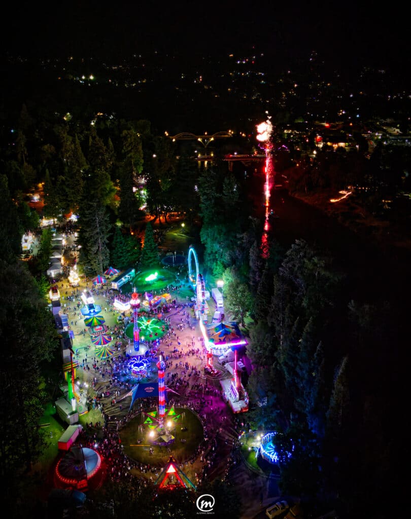 Aerial night view of a glowing carnival and a firework over the river at Boatnik 2024 in Grants Pass, Oregon.