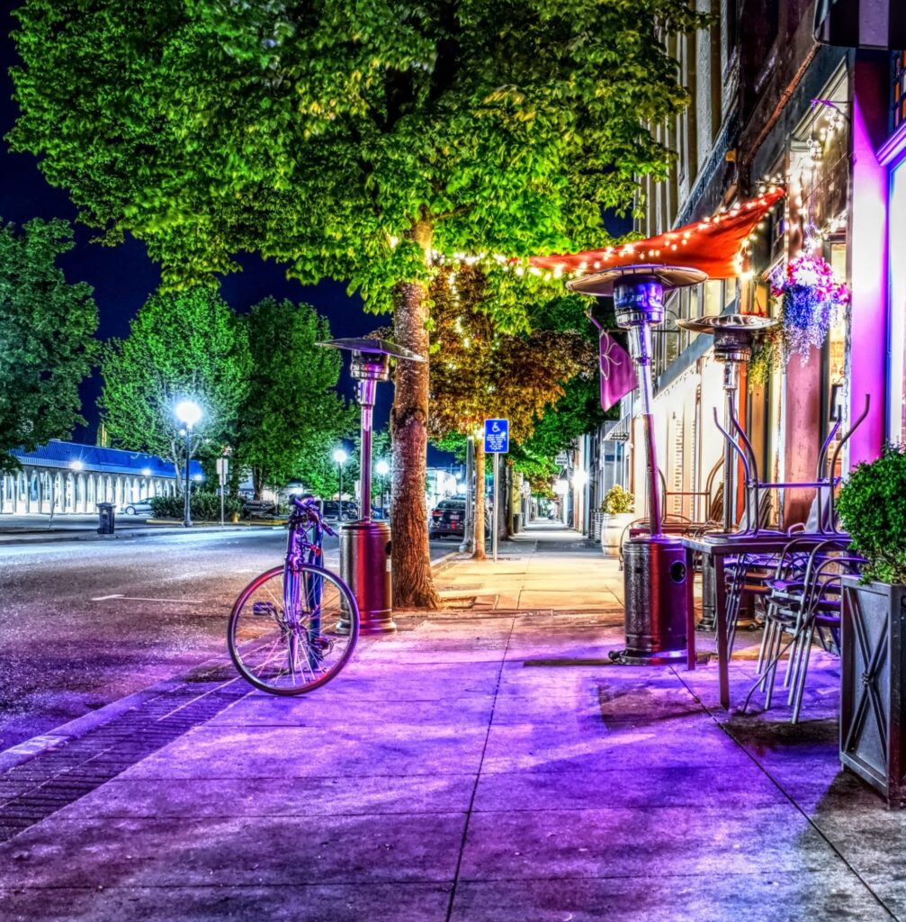 Vibrant night sidewalk in downtown Grants Pass with a bicycle, patio heaters, trees, and purple lighting.