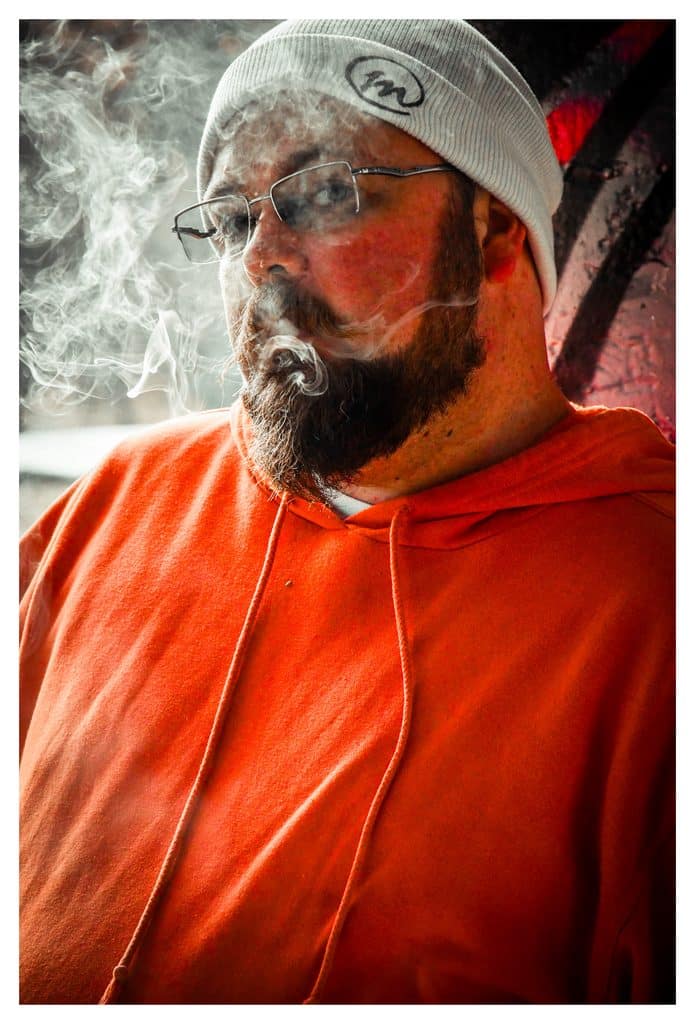 Bearded man in an orange hoodie and white beanie exhaling a thick cloud of smoke outdoors.