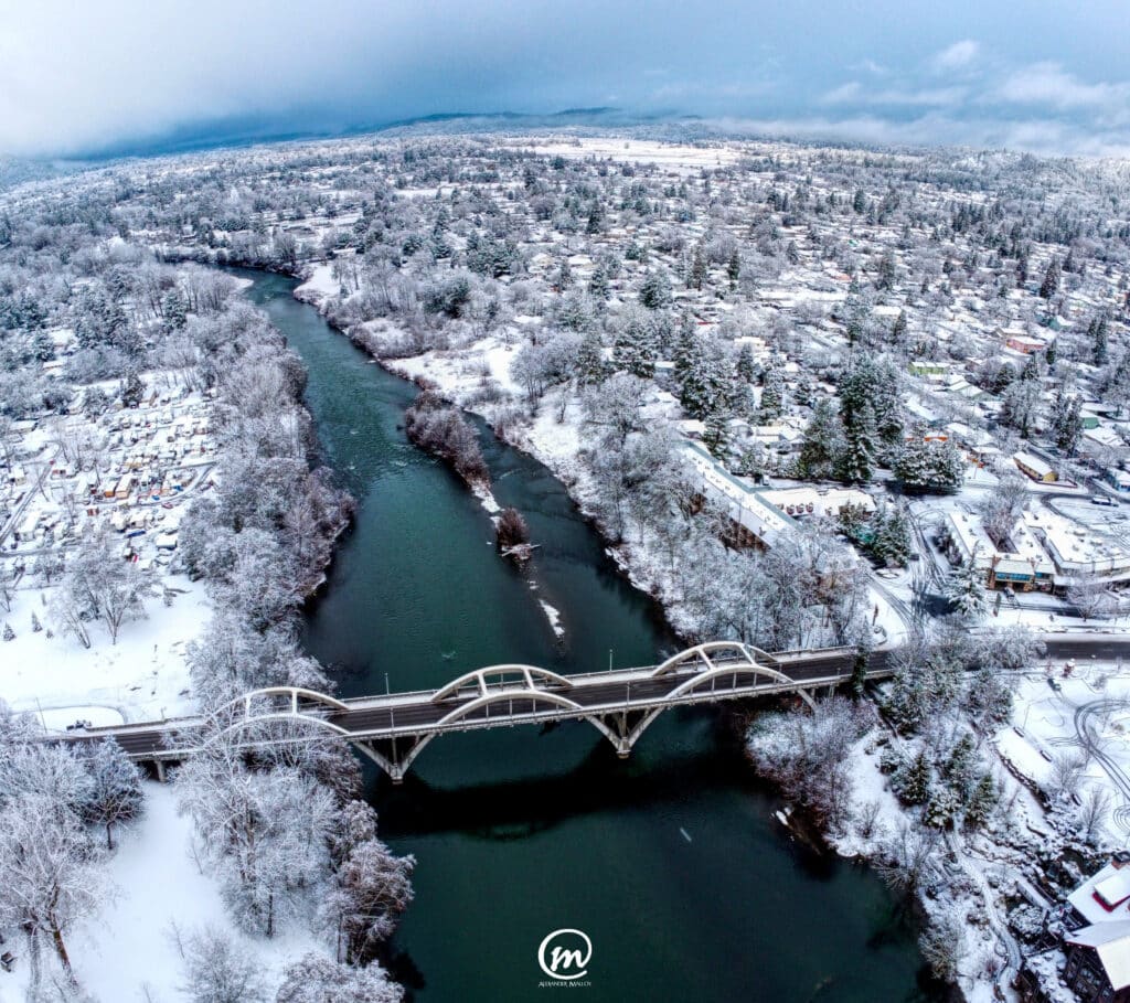 Aerial view of a snow-covered town and an arched bridge spanning a winding river under a cloudy sky.